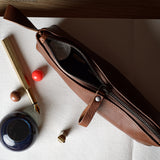A brown leather zippered pencil case or pouch lies open on a white sheet of paper, surrounded by stationery props including a gold pen, ink bottle, and small red and brown objects, on a light wood surface.