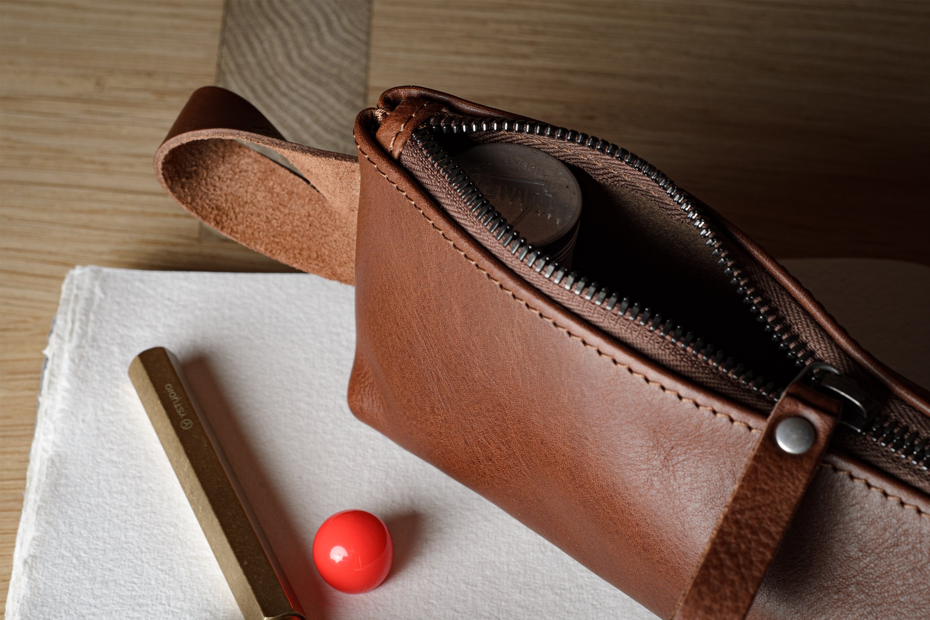 A close-up of a brown leather zippered pouch or cosmetic bag lying on a white cloth, with a gold pen and a small red sphere nearby on a wooden surface.