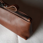 A close-up of a brown leather zippered pouch or case resting on a textured white fabric surface.