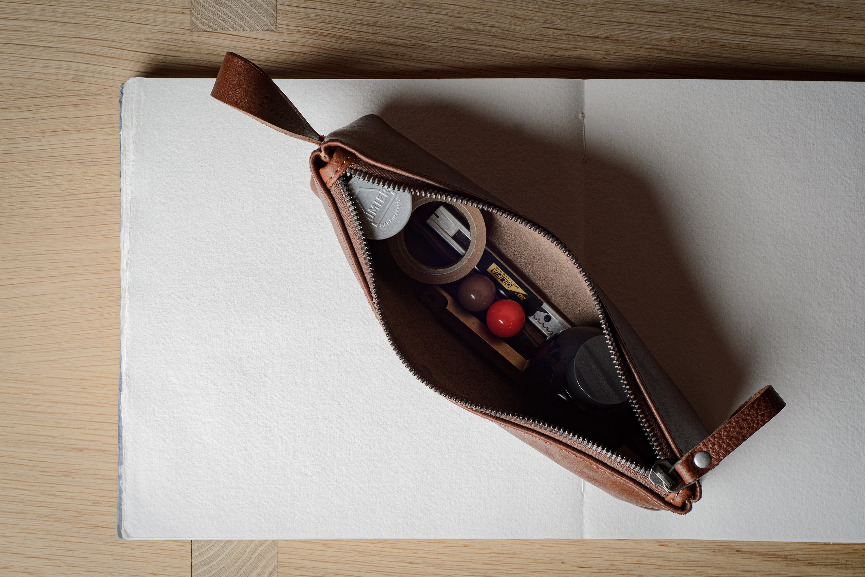 A brown leather zippered pouch with a pull tab, shown open on a white notebook over a light wood surface. The pouch contains various small cosmetic items.