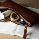 A brown leather zippered pencil case or pouch, partially open to reveal stationery items inside, resting on an open white notebook over a light wood surface.