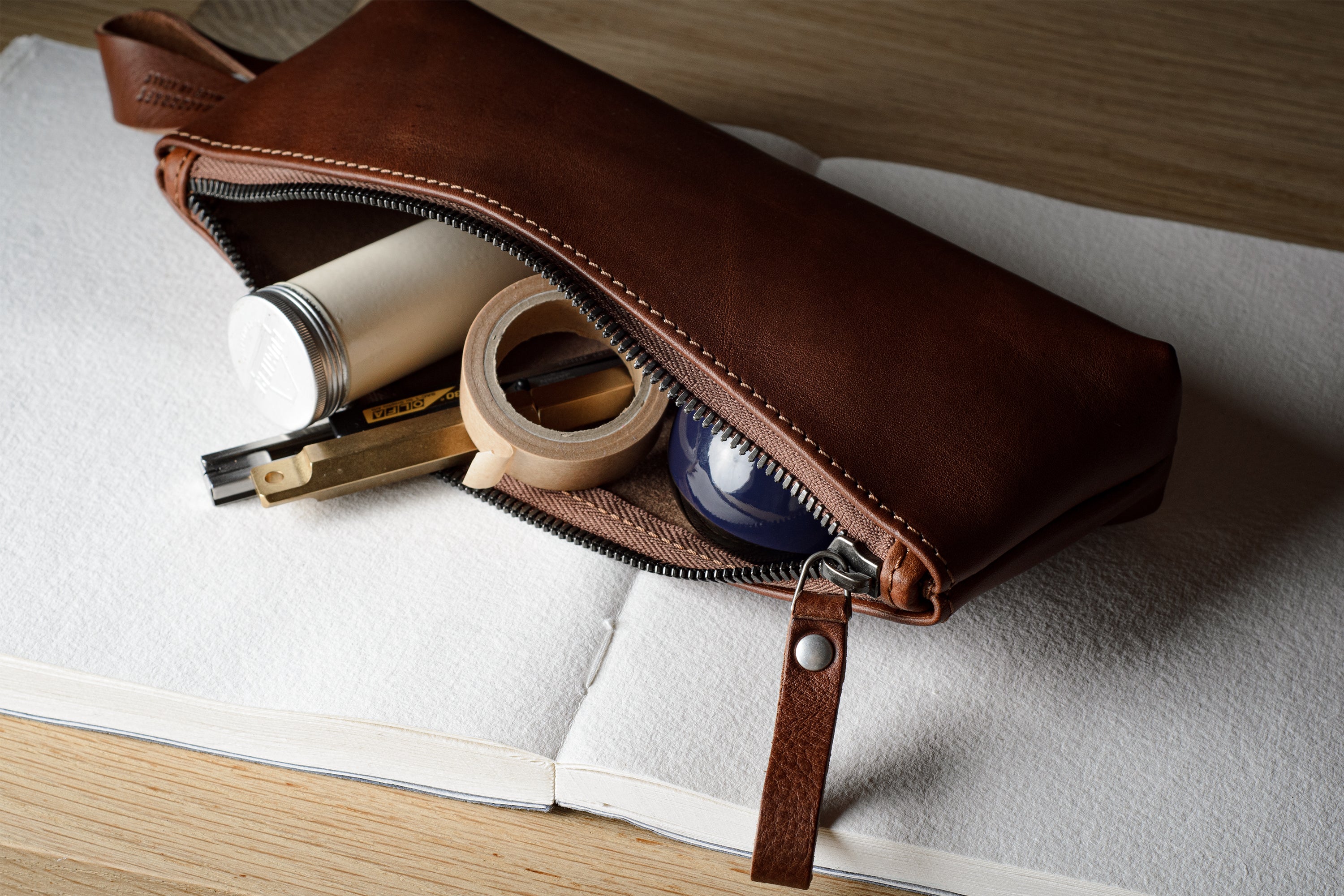 A brown leather zippered pencil case or pouch, partially open to reveal stationery items inside, resting on an open white notebook over a light wood surface.