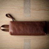 A brown leather zip pouch with a wrist strap, laid flat on a light wood grain surface.