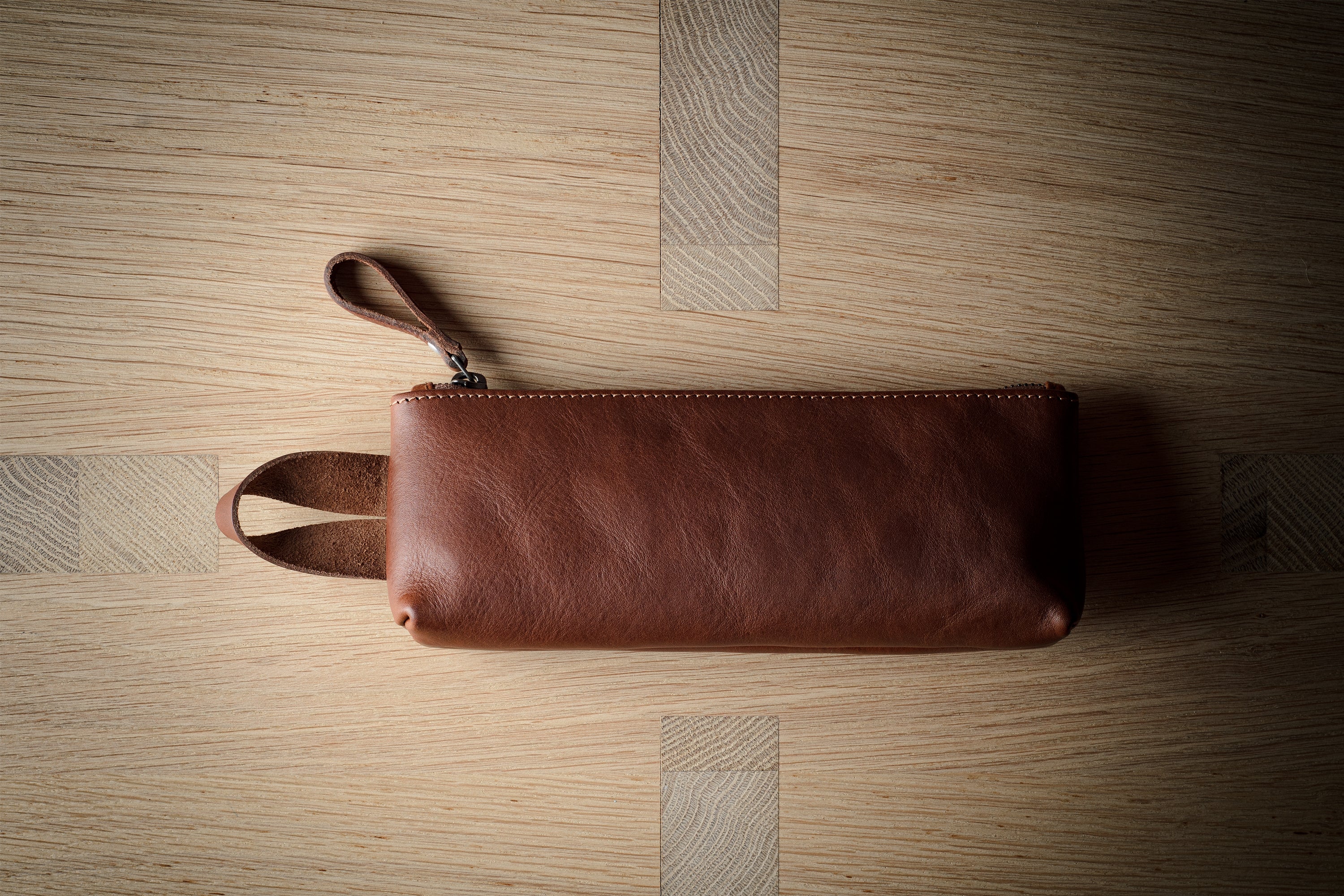 A brown leather zip pouch with a wrist strap, laid flat on a light wood grain surface.