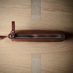 A brown leather zippered pouch with a leather pull tab, photographed from above on a light wood grain surface.