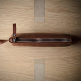 A brown leather zippered pouch with a leather pull tab, photographed from above on a light wood grain surface.
