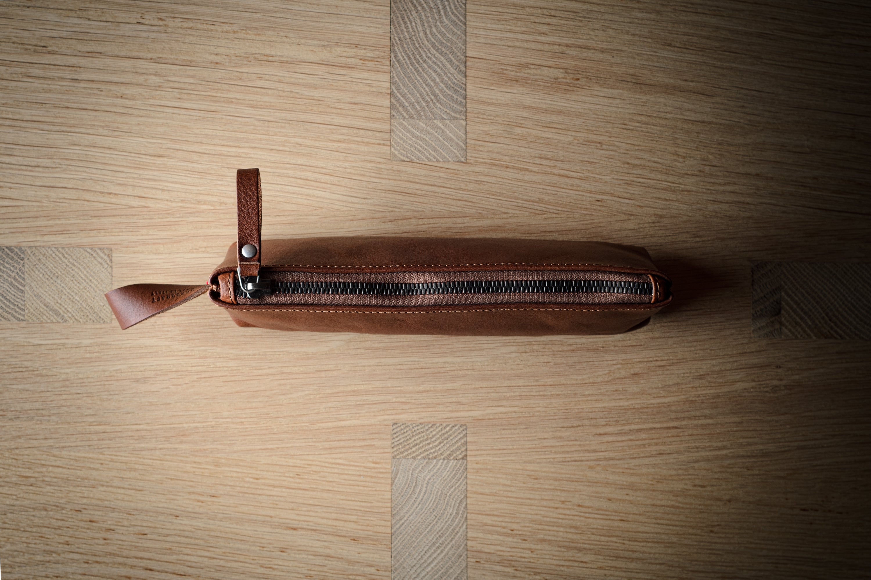 A brown leather zippered pouch with a leather pull tab, photographed from above on a light wood grain surface.