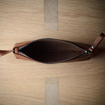 An open brown leather zip pouch with a leather pull tab, photographed from directly above on a light wood grain surface.