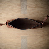 An open brown leather zip pouch with a leather pull tab, photographed from directly above on a light wood grain surface.