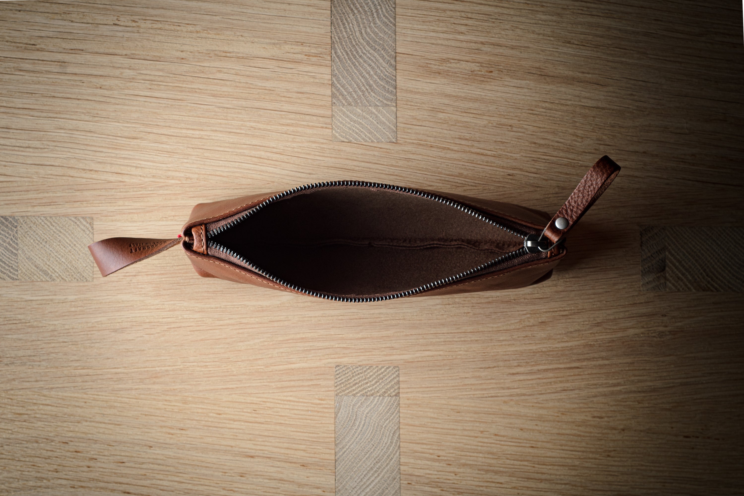An open brown leather zip pouch with a leather pull tab, photographed from directly above on a light wood grain surface.