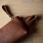 A brown leather clutch or sleeve with a wrist strap, partially unrolled on a light wood grain surface.