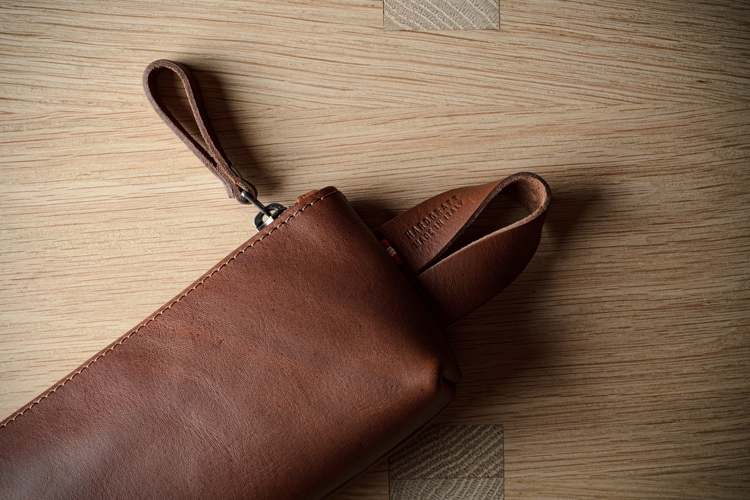 A brown leather clutch or sleeve with a wrist strap, partially unrolled on a light wood grain surface.