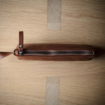 A brown leather zippered pouch with a leather pull tab, laid horizontally on a light wood grain background.