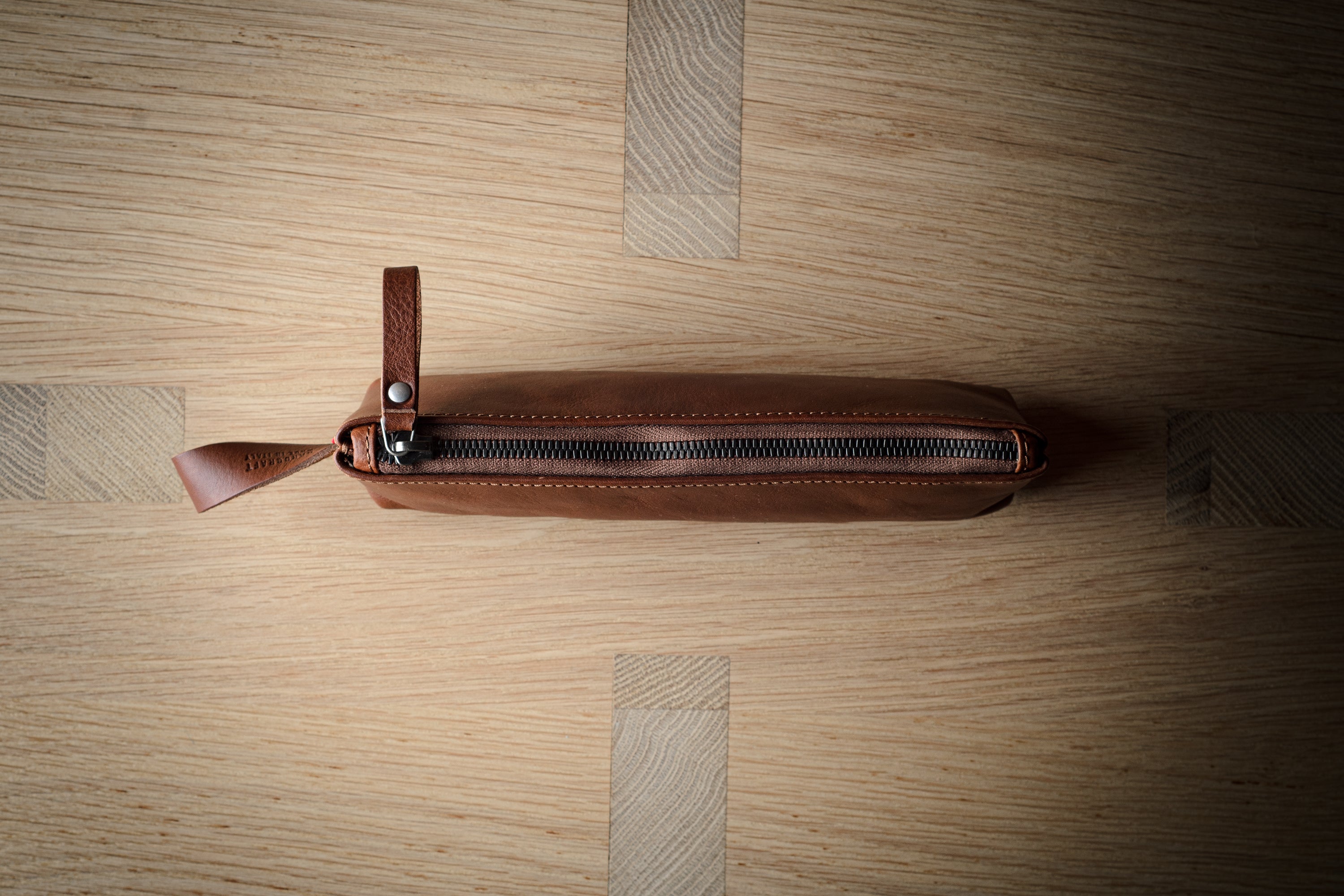 A brown leather zippered pouch with a leather pull tab, laid horizontally on a light wood grain background.