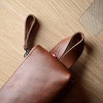 A close-up of a brown leather pouch or sleeve with a wrist strap, placed on a light wood floor.