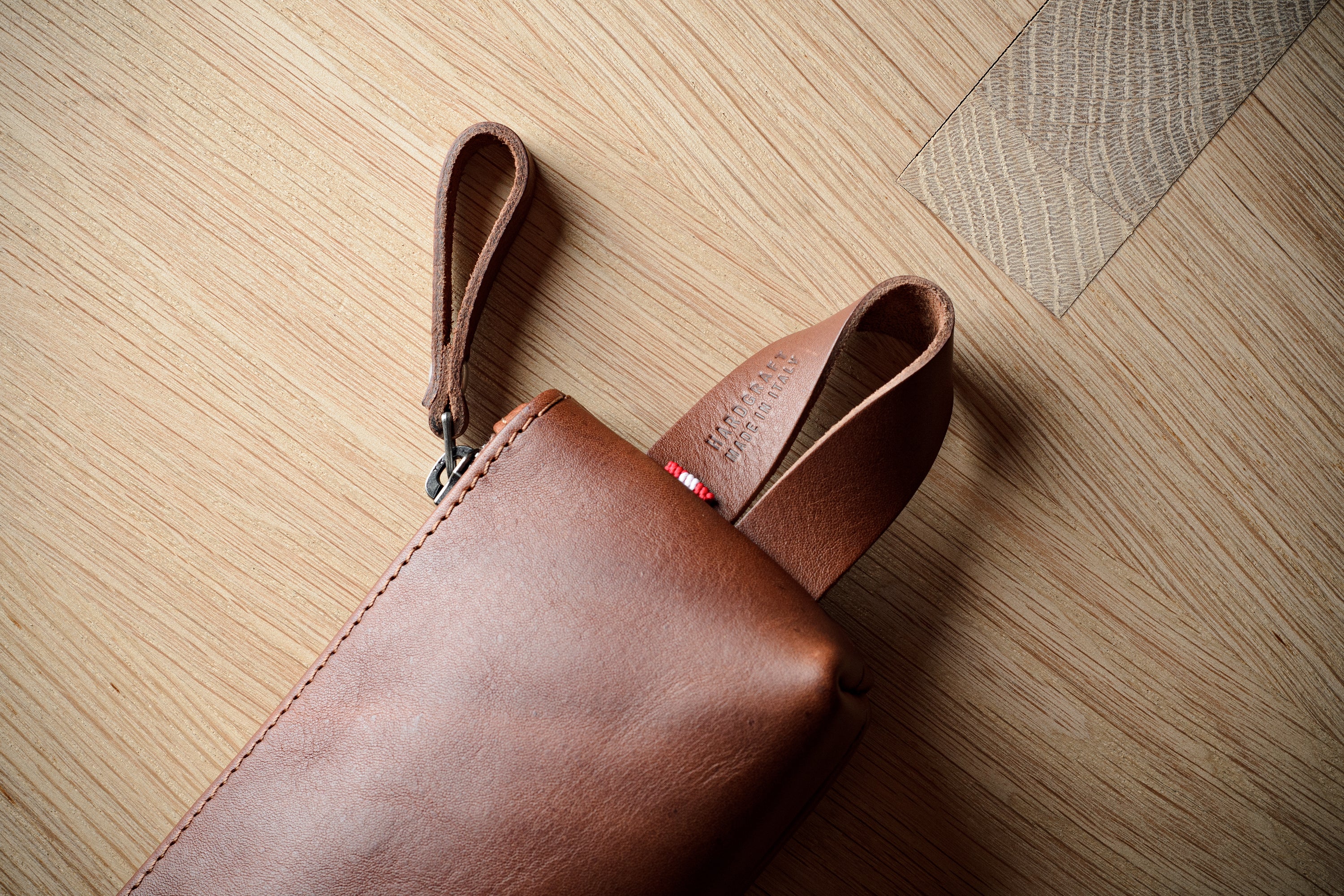 A close-up of a brown leather pouch or sleeve with a wrist strap, placed on a light wood floor.