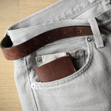 A close-up of light gray denim jeans with a brown leather belt tucked into the back pocket, with a matching brown leather card holder or wallet also visible in the pocket, set against a light wood background.
