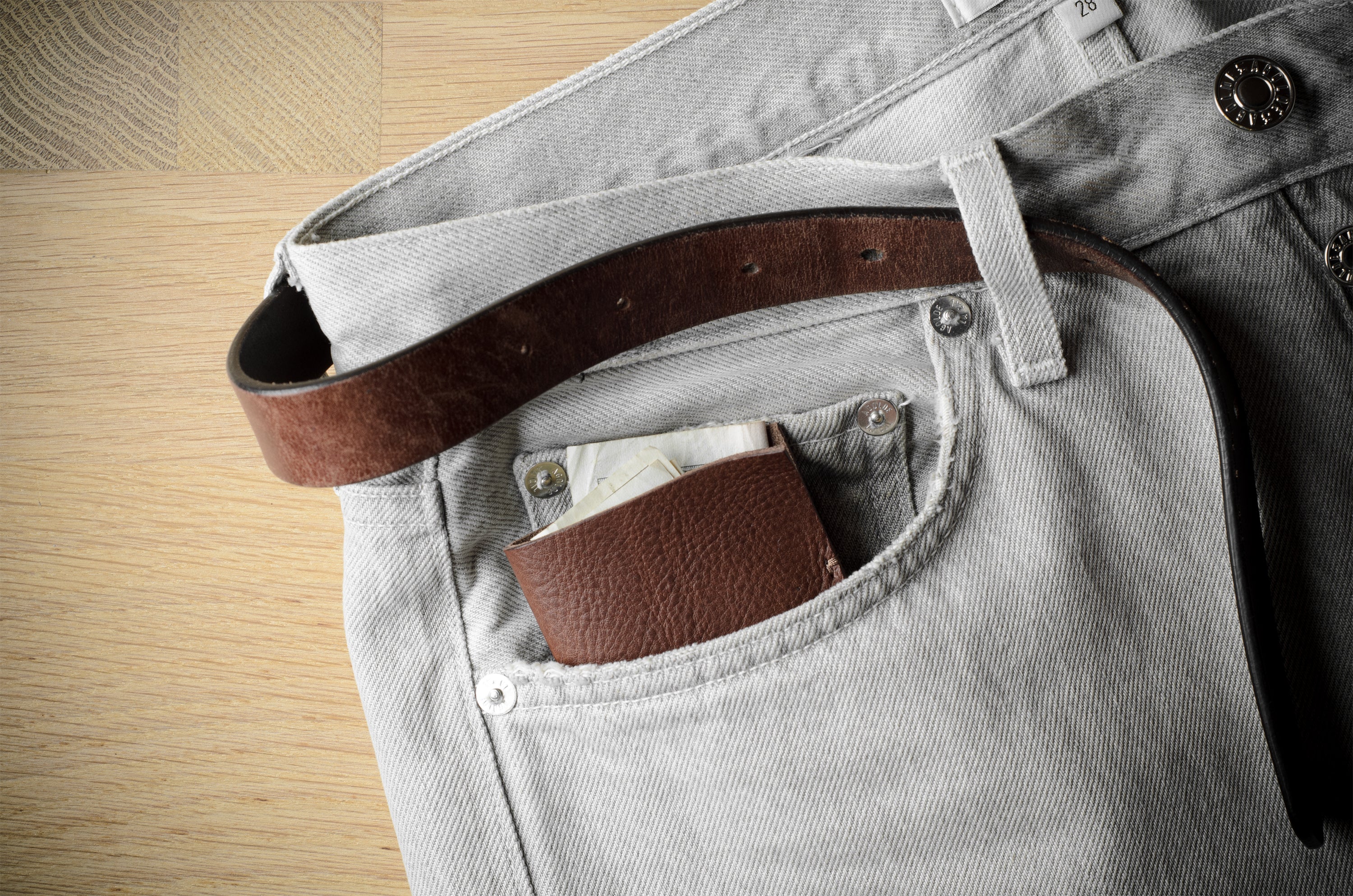 A close-up of light gray denim jeans with a brown leather belt tucked into the back pocket, with a matching brown leather card holder or wallet also visible in the pocket, set against a light wood background.