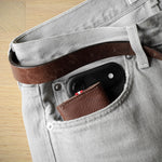 Close-up of a light gray denim jeans pocket holding a brown leather belt and a small brown leather card holder, with a black smartphone tucked into the pocket, set against a light wood floor background.