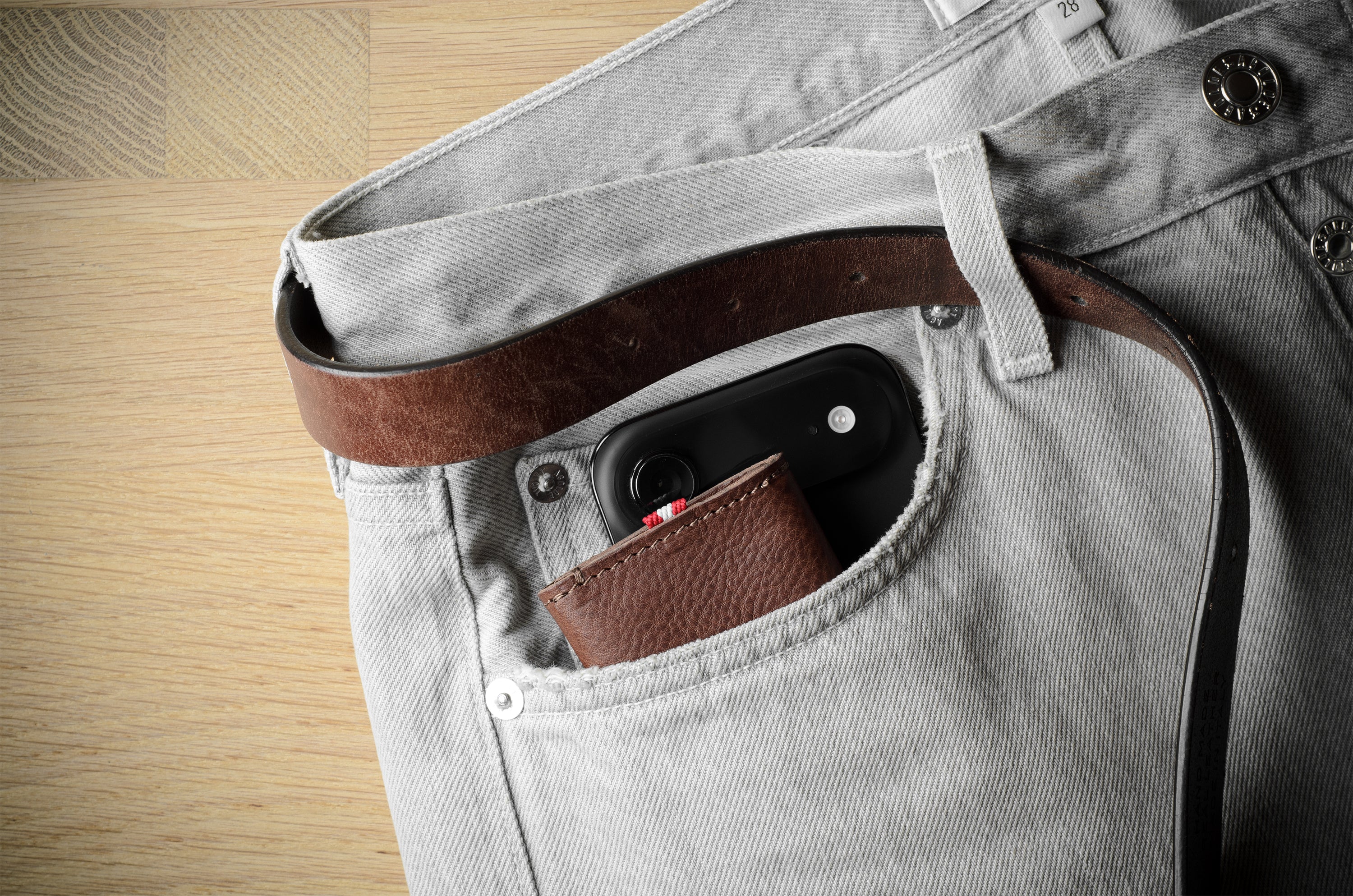 Close-up of a light gray denim jeans pocket holding a brown leather belt and a small brown leather card holder, with a black smartphone tucked into the pocket, set against a light wood floor background.