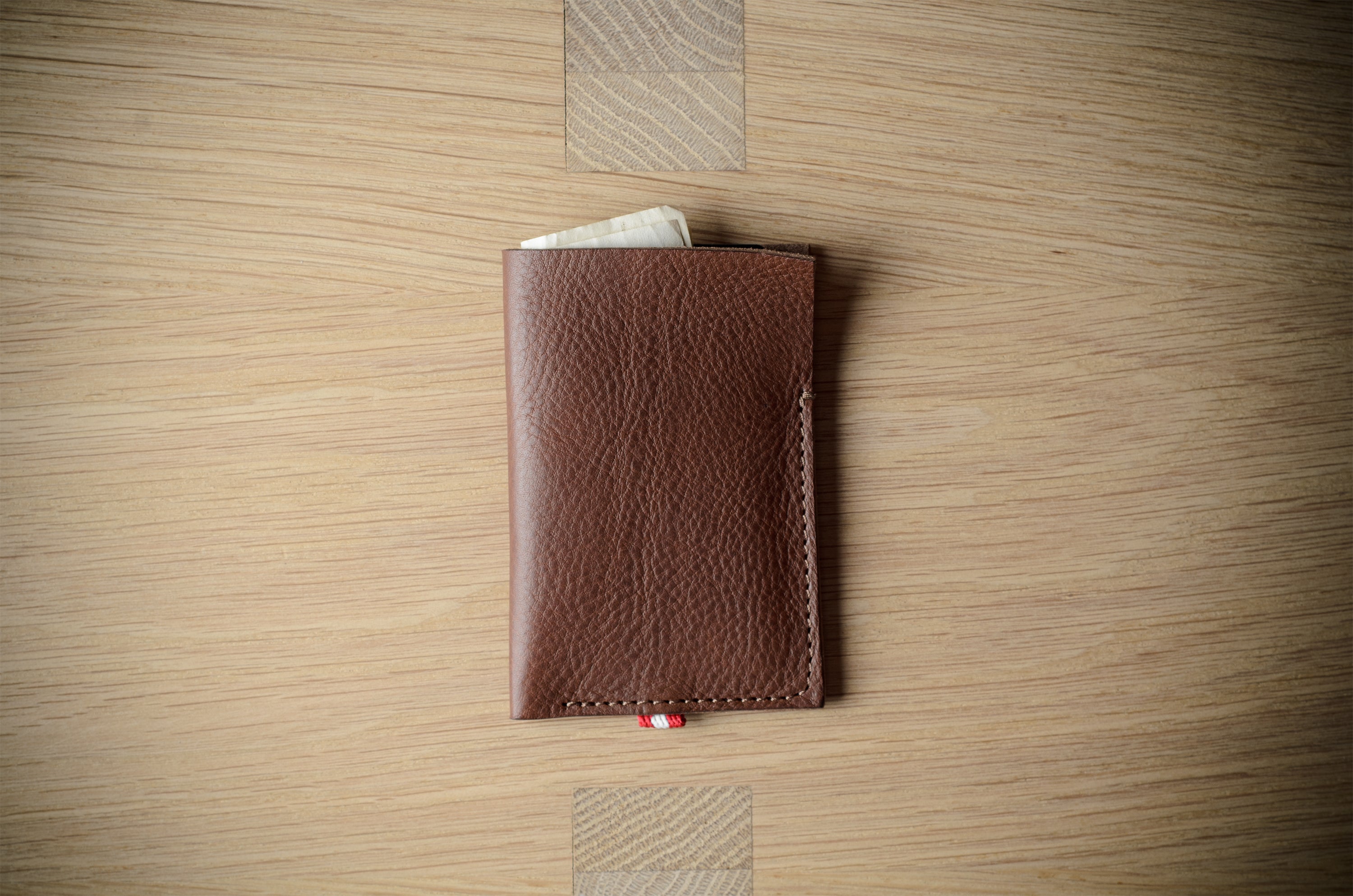 A brown textured leather card holder wallet with visible stitching and a small red-and-white cord detail, photographed from above on a light wood grain surface.