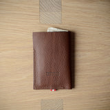 A brown textured leather card holder wallet with a small red and white striped ribbon bookmark, displayed on a light wood grain surface.