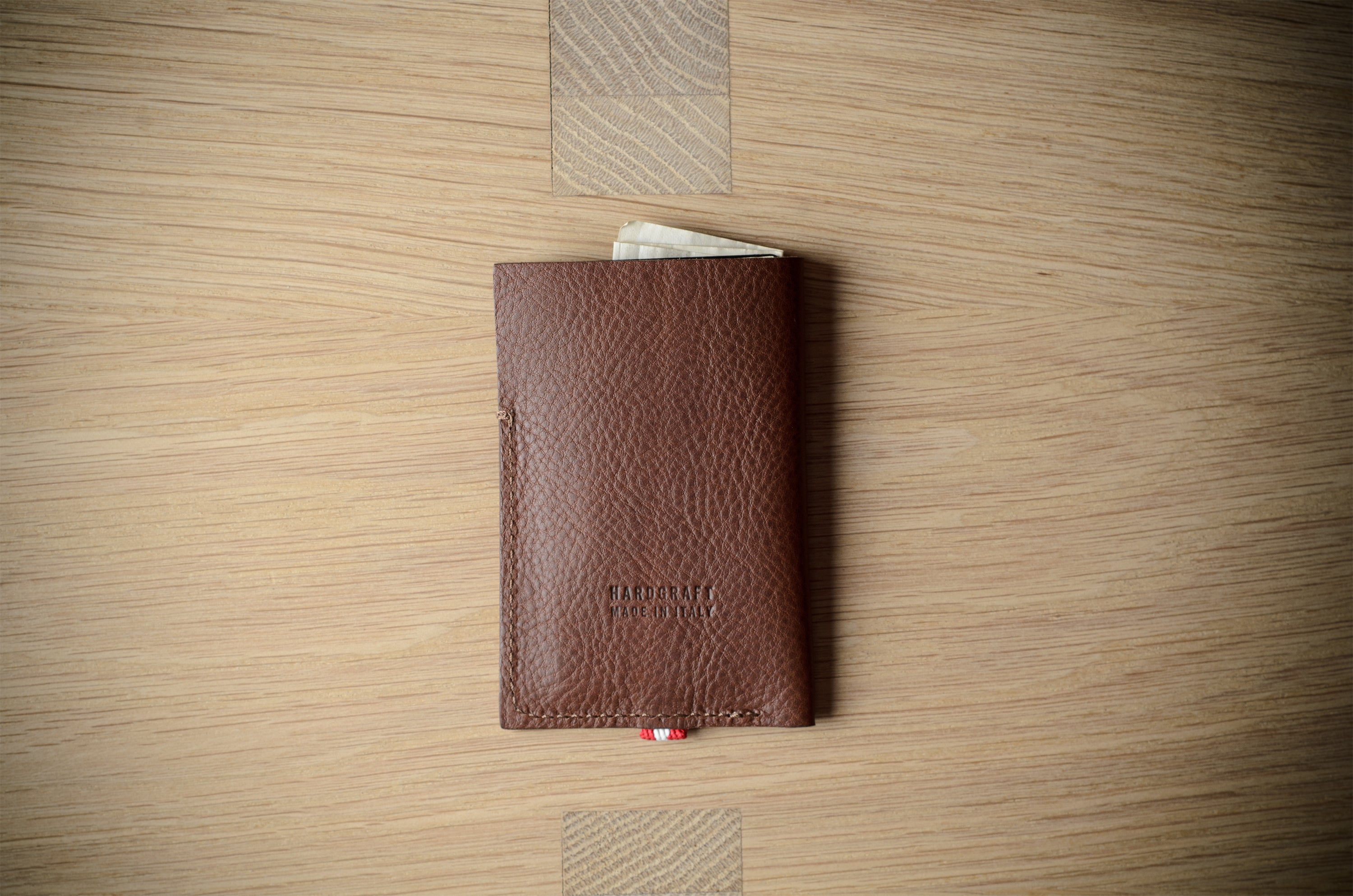 A brown textured leather card holder wallet with a small red and white striped ribbon bookmark, displayed on a light wood grain surface.