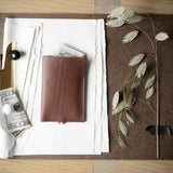 A brown leather card holder wallet with a small red tab, displayed on a brown leather folder with white paper, a gold clip, and dried leaves on a wooden table.