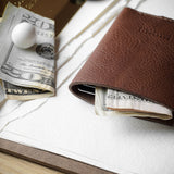 A brown leather card holder or slim wallet with a few dollar bills and a small white marble inside, styled on a white textured paper surface with a wooden table edge visible.