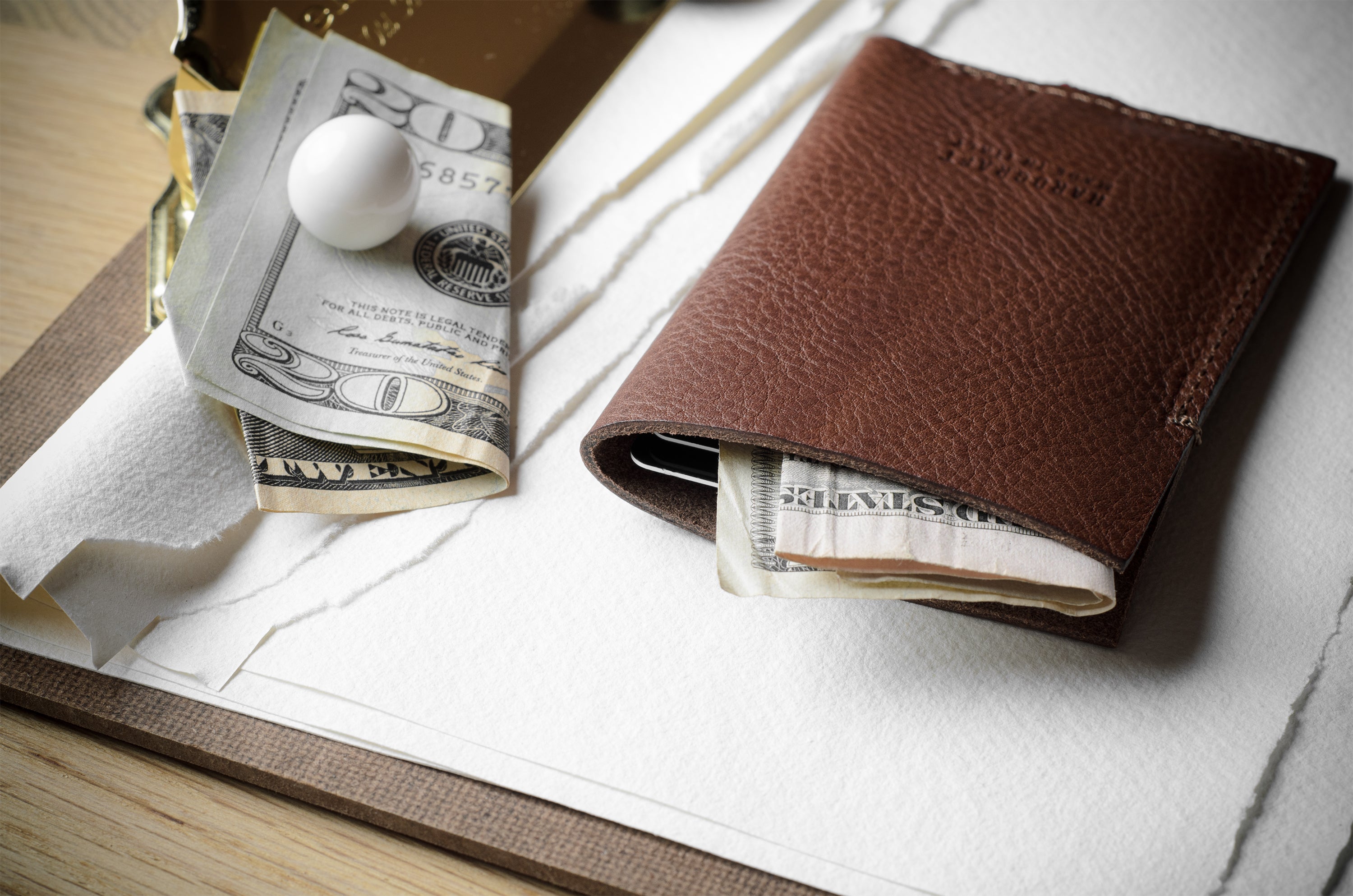 A brown leather card holder or slim wallet with a few dollar bills and a small white marble inside, styled on a white textured paper surface with a wooden table edge visible.