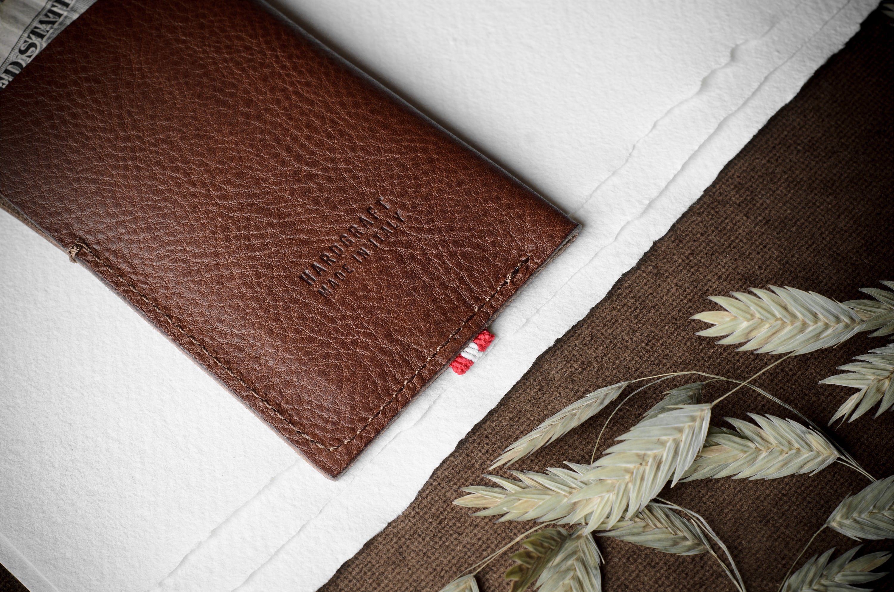 A brown textured leather card holder or wallet with 'HARDGRAFT MADE IN ITALY' embossed on the front, styled on a white paper background with a brown burlap section and dried oat stems.