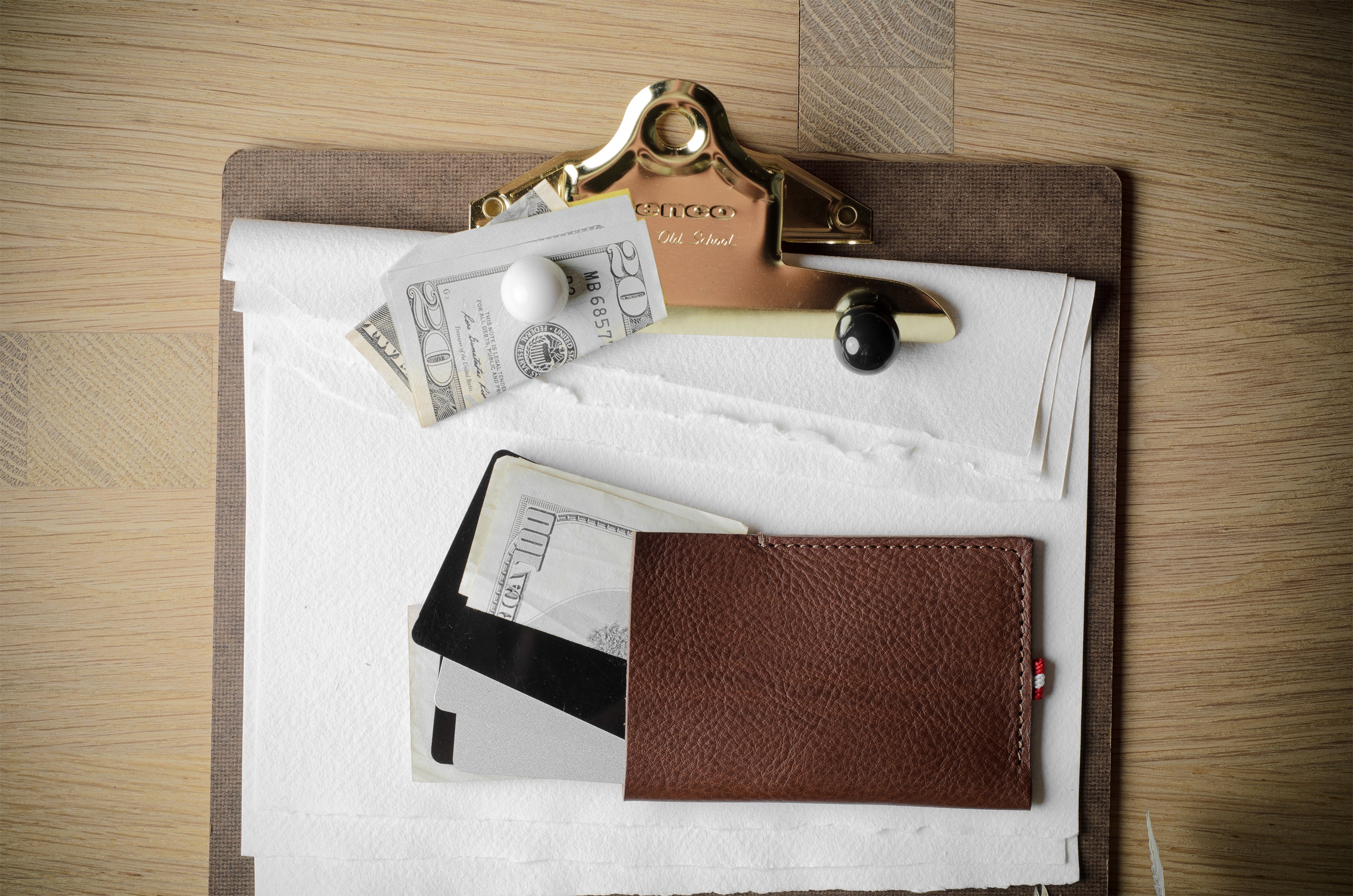 A brown leather wallet or card holder pouch lies on a white sheet of paper atop a clipboard, alongside cash, a credit card, and a gold-toned binder clip on a wood surface.