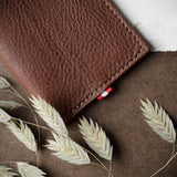 A brown textured leather card wallet, shown closed on a brown burlap-like surface with dried oat stalks and a white paper corner visible.