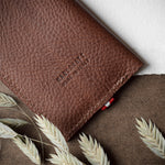 A brown textured leather card case or wallet with 'HARDGRAFT MADE IN ITALY' embossed on the front, styled flat-lay with dried oat stalks on a brown burlap surface and white paper in the background.
