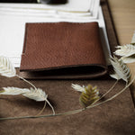 A brown textured leather bifold wallet placed on a brown fabric surface, styled with dried white wheat stems and surrounded by papers on a wooden desk.