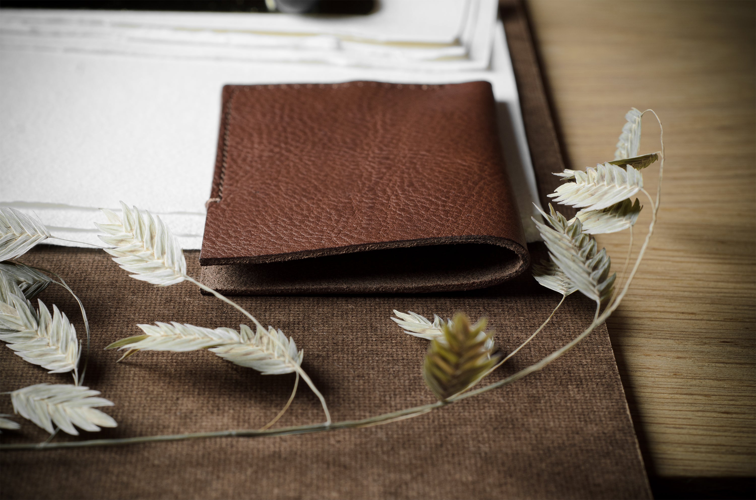 A brown textured leather bifold wallet placed on a brown fabric surface, styled with dried white wheat stems and surrounded by papers on a wooden desk.