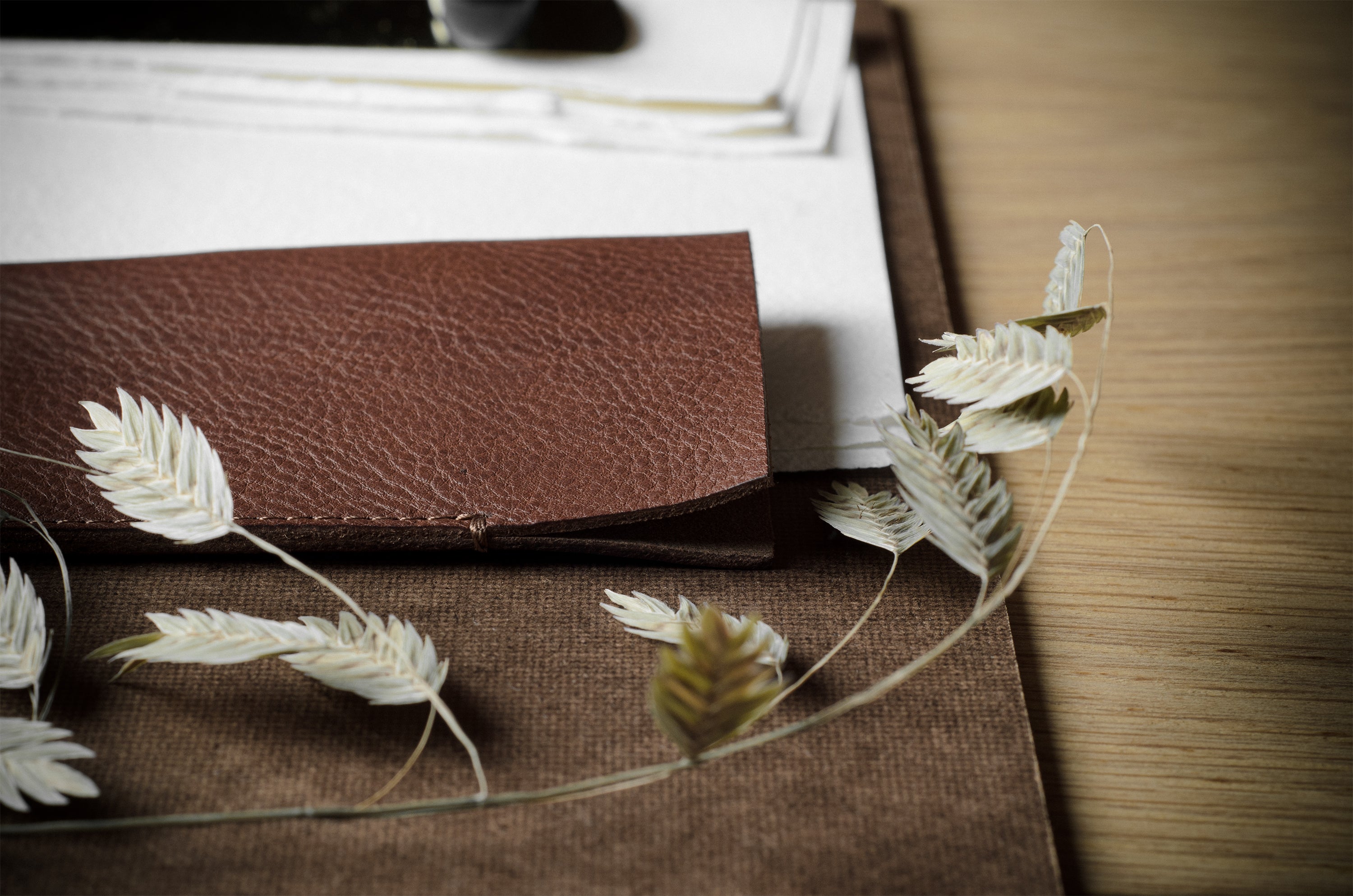 A close-up of a brown leather card case resting on a brown fabric surface, with dried white wheat-like leaves draped across it on a wooden tabletop.