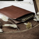 A brown leather card holder wallet with card slots, shown open with cards partially visible, styled on a brown fabric surface with dried oat grass sprigs.