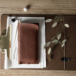 A brown textured leather iphone case placed on a brown clipboard with white papers, a brass clip, and dried plant stems on a light wood tabletop.