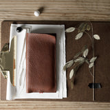 A brown textured leather iphone case placed on a brown clipboard with white papers, a brass clip, and dried plant stems on a light wood tabletop.