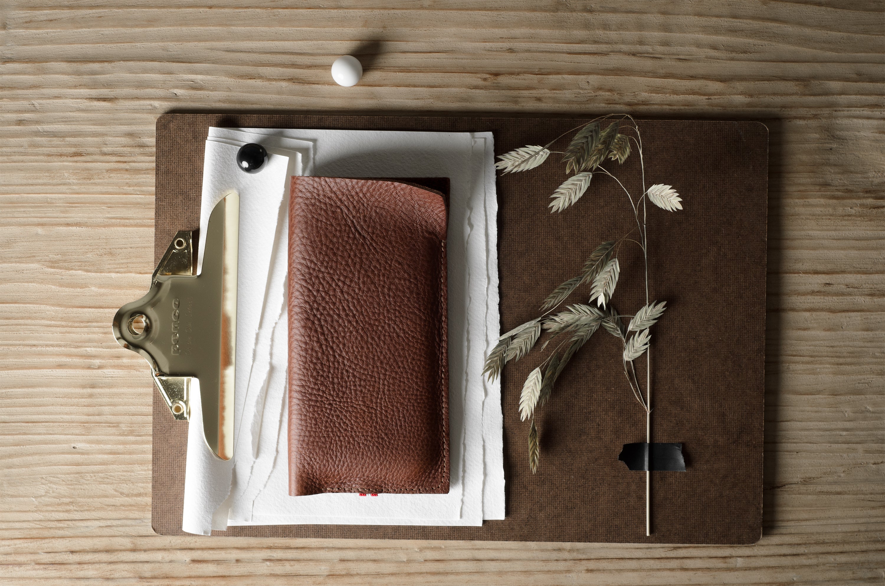 A brown textured leather iphone case placed on a brown clipboard with white papers, a brass clip, and dried plant stems on a light wood tabletop.