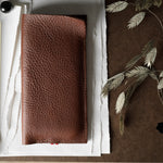 A brown textured leather iphone case laid flat on white paper, with a brass binder clip on the left and dried plant stems on a brown surface to the right.