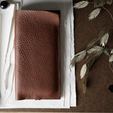 A brown textured leather iphone case laid flat on white paper, with a brass binder clip on the left and dried plant stems on a brown surface to the right.