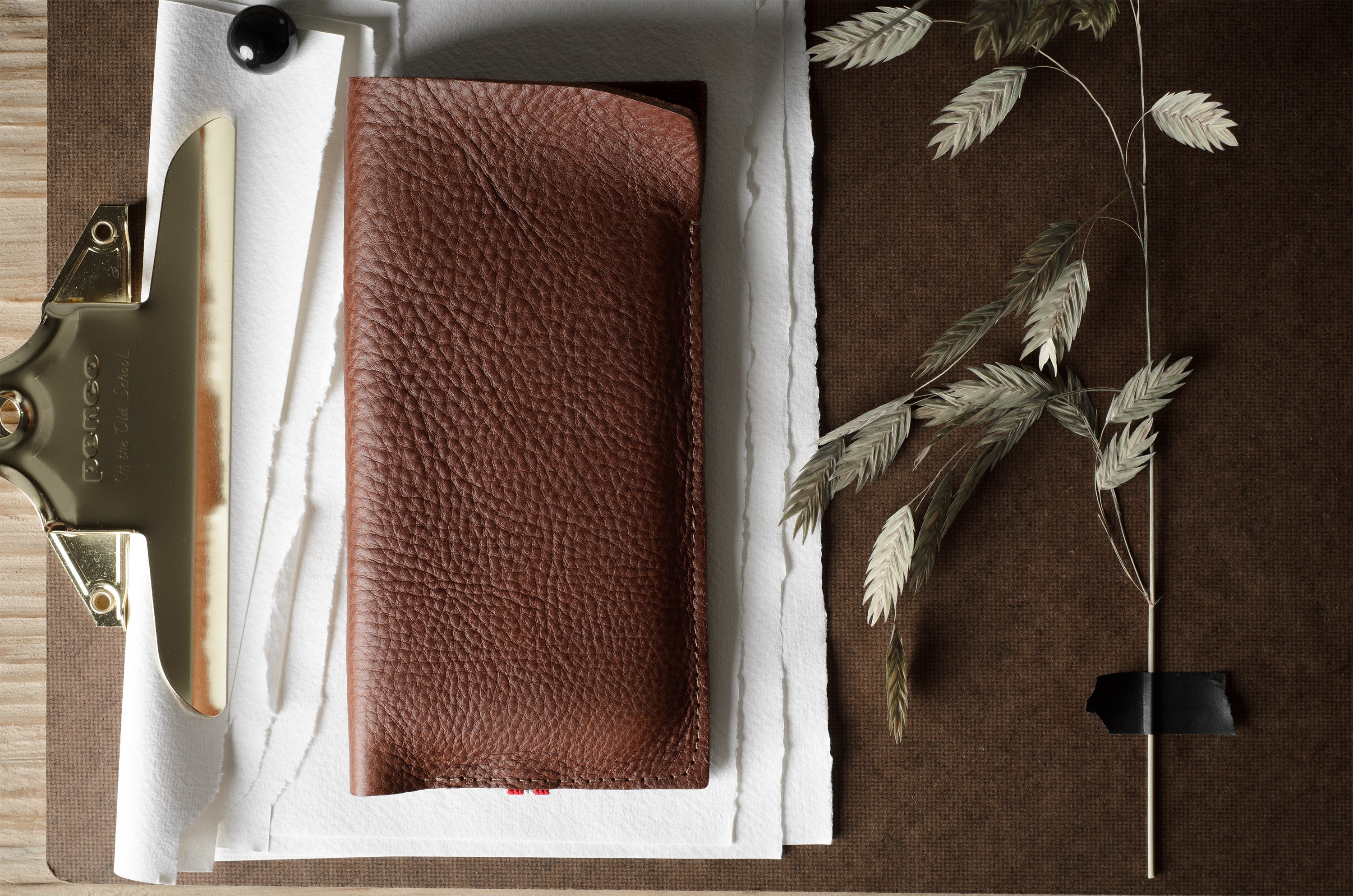 A brown textured leather iphone case laid flat on white paper, with a brass binder clip on the left and dried plant stems on a brown surface to the right.