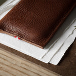 A close-up of a brown textured leather iphone case resting on white paper over a wood surface, with a small red thread detail visible along the edge.