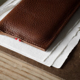 A close-up of a brown textured leather iphone case resting on white paper over a wood surface, with a small red thread detail visible along the edge.