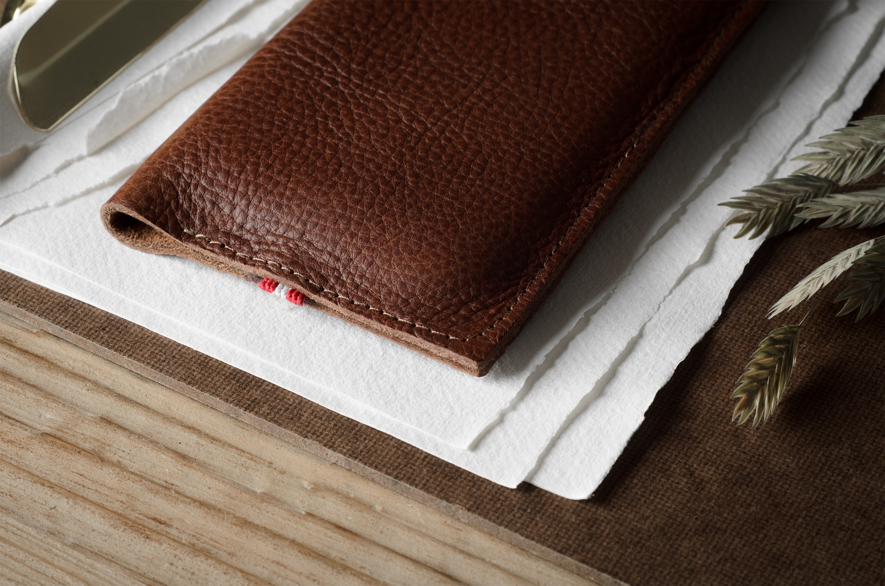 A close-up of a brown textured leather iphone case resting on white paper over a wood surface, with a small red thread detail visible along the edge.