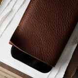 A close-up of a brown textured leather iphone case partially covering a black smartphone, styled on a white textured paper surface with dried grass and a gold object in the background.