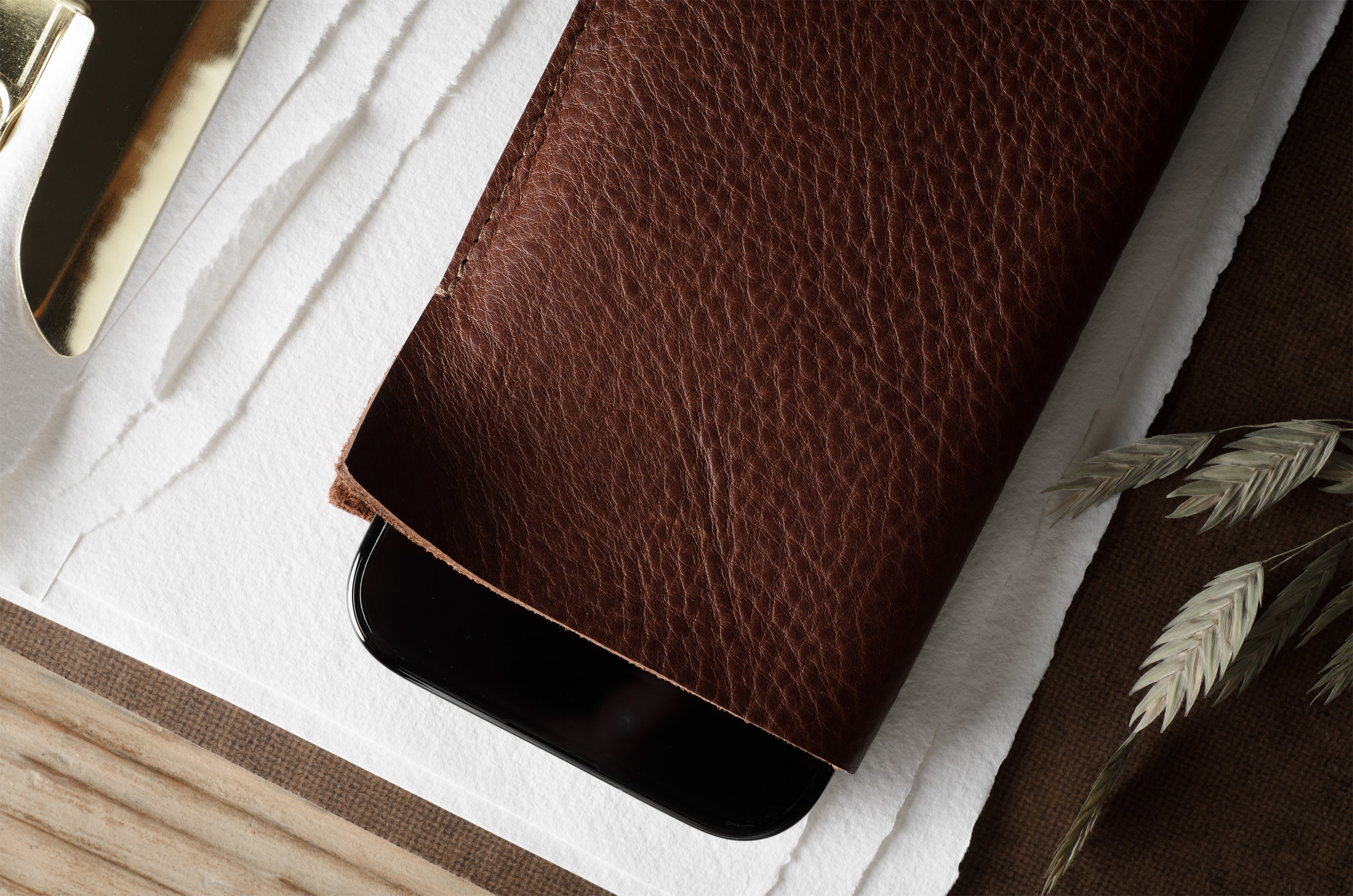 A close-up of a brown textured leather iphone case partially covering a black smartphone, styled on a white textured paper surface with dried grass and a gold object in the background.