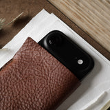 A close-up of a black smartphone partially tucked into a brown textured leather iphone case, placed on white paper over a wood-grain surface with dried grasses in the corner.